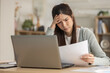 © Ratirat - Stressed Asian woman feeling worried about bill and debt. She has financial problem, sitting with laptop at home and holding paper with confused look