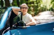 © standret - Middle-aged woman with short gray hair and glasses leans out of a blue car window, smiling in a sunlit forested area along a winding road