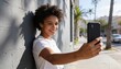 © Future Vision - Leaning woman wearing white T-shirt, holding black smartphone and smiling on sunlit city sidewalk