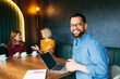 © djile - Confident businessman smiling at camera while sitting with laptop and coworkers during relaxed business meeting in modern cafe