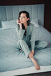 © deagreez - Young woman in green pajamas sitting on bed looking thoughtful in a cozy morning bedroom lifestyle portrait