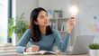 © ArtFocus Studio - A young Asian woman holding a glowing lightbulb while sitting at her desk with a laptop and books, representing creativity, innovation, and successful problem-solving in a bright office.