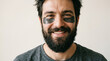 © Cliff - Close-up portrait of a happy, brutal young man with a thick beard wearing cosmetic under-eye patches, smiling at the camera. Clean skincare and self-care concept for men