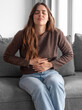 © luciano - Young Woman Sitting on Sofa Holding Stomach in Pain Showing Abdominal Discomfort and Distress