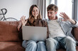 © luciano - Young Couple Waving During Video Call On Laptop While Sitting Together On Couch At Home