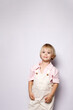 © Krystsina - Cute blonde toddler boy standing and smiling in studio. Happy relaxed little child looking at camera. Sweet kid wearing beige overalls and pink shirt. Vertical portrait isolated on white background