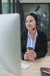 © MINAE - Young Asian businesswoman at desk wearing headset, smiling while providing online customer support and telemarketing via desktop computer, holding a coffee cup and working remotely