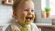 © Kleber - Happy baby covered in green food mess enjoying mealtime messy eating fun