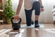 © Nanci - Mature woman in activewear doing a kneeling kettlebell lunge in her living room, maintaining strength training and a healthy, sustainable fitness routine for wellness and mobility