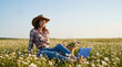 © Дина Етова - A young woman in a cowboy suit is sitting in a clearing among daisies and working on a laptop remotely. Freelancer out of town.