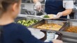 © Olga Ко - School cafeteria food service concept: Over-the-shoulder view of a child waiting in line while a worker serves a healthy hot lunch on a plate