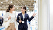 © Yka - 展示会で女性客を笑顔でブースへ案内するビジネスウーマン