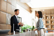 © fizkes - Male receptionist assisting to female guest at front desk, processing check-in, answering questions, explaining booking reservation detail, provide professional hospitality services in hotel reception