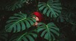 © yay - Young woman with vibrant red hair partially obscured by lush green monstera deliciosa leaves in a dark botanical setting
