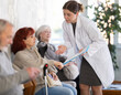 © JackF - Polite young nurse or doctor talking to mature woman patient standing in light waiting room of medical clinic, with her back to visitors sitting in chairs