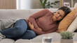 © ArtFocus Studio - Young woman lying on couch holding her stomach in pain, suffering from abdominal discomfort, menstrual cramps, or food poisoning at home