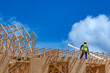 © Andrei Liashchou - Construction worker standing on the top plate of a wooden house frame and carrying a wood board near roof trusses under a blue sky with clouds, USA