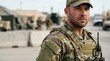© xuxu - Focused male soldier in camouflage uniform and tactical vest with military base in the background