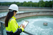 © Viktor - Asian woman engineer in hard hat and hi-vis jacket checks water treatment plant facility on clipboard. She records data on a large sewage treatment tank, monitoring water quality during inspection.