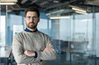 © Liubomir - Confident bearded businessman wearing eyeglasses and a casual sweater, standing with crossed arms in a contemporary corporate office, portraying professionalism and leadership