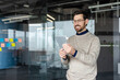 © Liubomir - Smiling man standing in a modern office, wearing glasses and a sweater, holding a digital tablet and working while looking at the screen with a happy expression