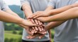 © Alice M - Multiple hands stacked together in unity outdoors. People in gray shirts demonstrate teamwork and collaboration against a scenic green landscape, symbolizing group commitment and support.
