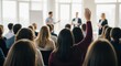 © Alice M - A group of diverse people in a bright room with large windows, listening to speakers. One person raises their hand to participate in this business meeting, conference, or training session.