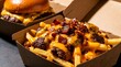 © Studio Fiuza - Close up of delicious loaded french fries with melted cheddar cheese, crispy bacon, and meat in a takeout cardboard box. A blurry cheeseburger is visible in the background, perfect for fast food.