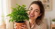 © Konstantin - Happy young woman holding a potted green houseplant toward the camera, smiling warmly in a bright home—joyful indoor gardening and plant care moment