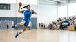 © Xenia - Dynamic female handball player jumping to throw the ball during a match in an indoor arena. Athletic woman in blue uniform performing a jump shot. Competitive sports and action concept