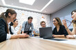 © bulentbaris - A diverse group of young professionals gather around a wooden conference table, engaged in a discussion led by a standing presenter.