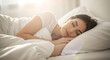 © LAB IMAGE - Woman sleeps peacefully in a soft white bed illuminated by gentle morning light streaming through a nearby window.