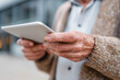 © Tina - Old hands hold tablet with care. Fingers show age and wisdom. Person focuses intently on screen. Urban backdrop softly blurred. Ideal for tech adoption themes. Perfect for senior learning or wellness