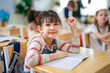 © Wanwajee - Happy young Asian student holding pencil and smiling at camera while sitting at classroom desk during elementary school lesson in modern multicultural environment.