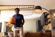 © wavebreak3 - African American man in 20s wearing navy T-shirt holding basketball on leather sofa in living room