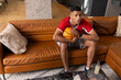 © wavebreak3 - African American man in 20s sitting on sofa at home holding basketball, wearing red shirt, watch