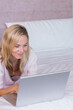 © wavebreak3 - Female reclining and leaning toward silver laptop on bed wearing light casual top