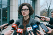 © AnnaStills - Portrait of young adult Caucasian businesswoman facing media microphones during press interview outside office building, representing public relations, news response, corporate messaging use