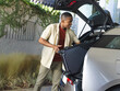 © wavebreak3 - African American man in beige shirt, olive pants lifting suitcase into silver hatchback on driveway