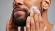 © ISVO - African American man applying facial cleanser to his beard while looking in the mirror, showcasing skincare routine and grooming habits in a neutral setting