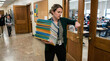 © logonv - Teacher holding stack of student notebooks while checking time at classroom door. Busy educator carrying homework assignments in school hallway. Academic work load and schedule concept.