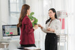 © Kritdanai - Office Collaboration: Two professional women engage in a friendly discussion while holding coffee cups in a modern office setting, with a computer displaying charts and a bookshelf in the background.