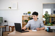 © Stella 16 - Smiling Asian Man Working from Home Writing Notes and using Laptop at Wooden Desk in Modern Home Office, remote work and lifestyle