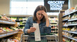 © Nataschen - Woman examines grocery receipt with a concerned expression in a supermarket, shelves filled with fresh produce in the background. For inflation, financial crisis and cost of living content