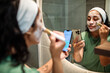 © Marko Geber - Young woman applying face mask while using smartphone in home bathroom