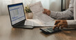 © onephoto - Accountant reviewing financial report and tax documents on laptop while using calculator for account analysis, real estate investment planning, expense management, and reporting workflow.