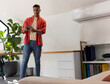 © wavebreak3 - Man standing and holding tablet, wearing red overshirt and jeans in home office with laptop, plant