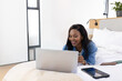 © wavebreak3 - African American woman in denim jacket lying in bedroom using laptop with tablet, notebook smiling