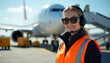 © Maryna - Woman airport worker wears headset and safety vest. She smiles in front of an airplane on tarmac. Ground staff manages aircraft operations and ensures smooth service.