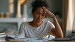 © AnastasiiaRozhmanova - Stressed Black woman surrounded by small business receipts, a spiral notebook, and an adding machine on a wood-grain desk, sole proprietor tax preparation, small business accounting, self-employment